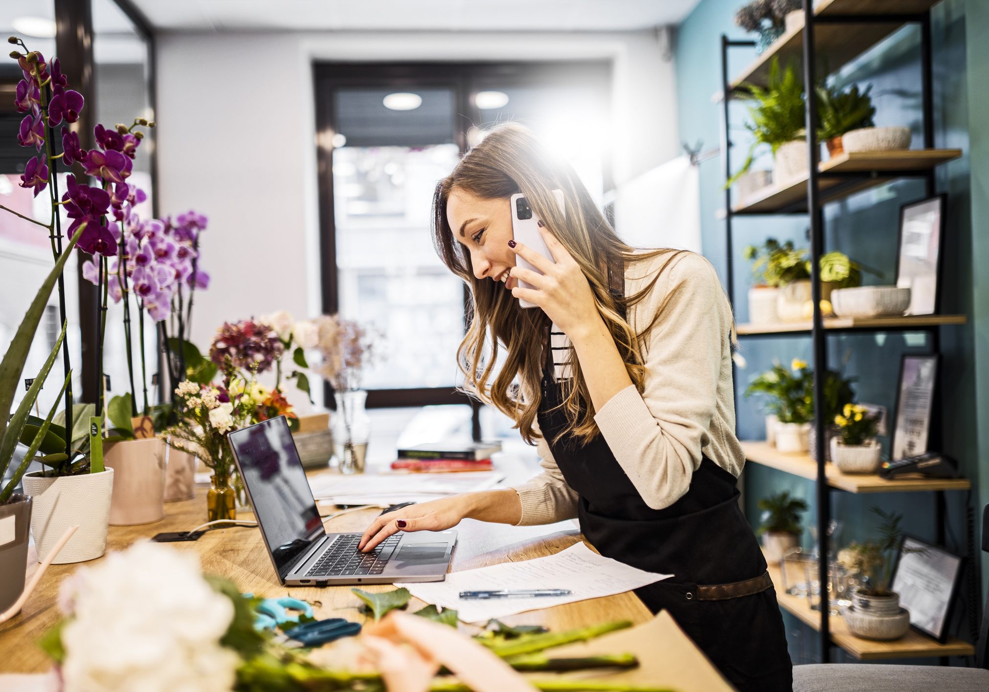 We got your order ,thank you for calling. Young florist using smartphone while using a laptop and making notes